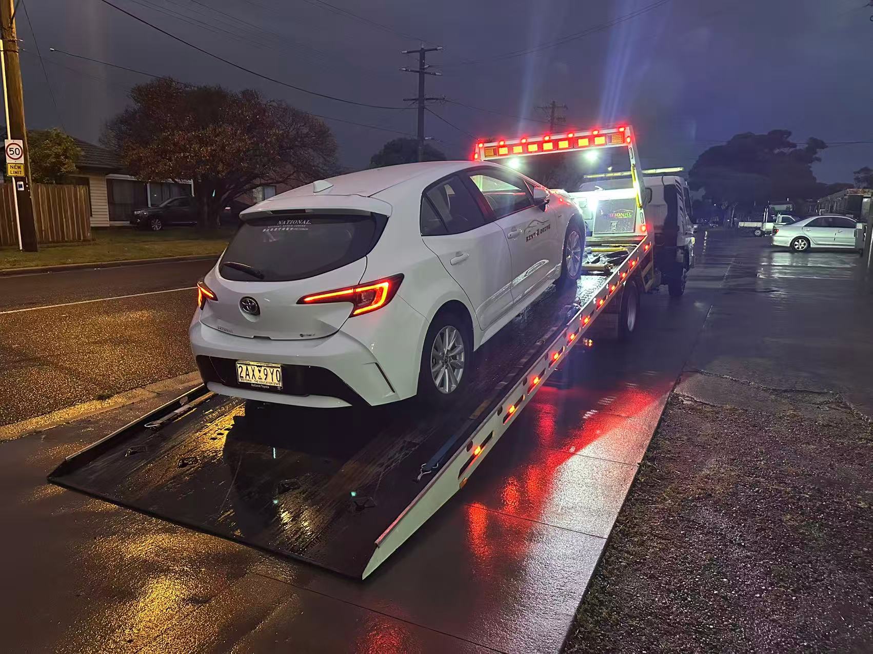 White hatchback being loaded onto a flatbed tow truck on a wet Melbourne street