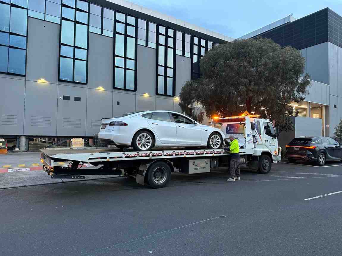 White electric sedan being loaded onto flatbed tow truck at dusk